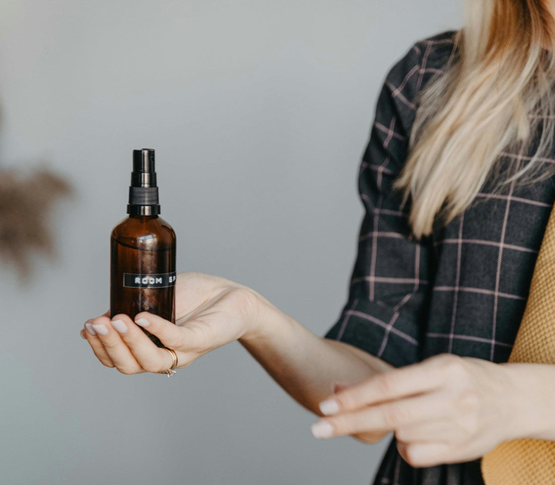 A person holding a small amber glass room spray bottle with a black pump top in their open hand.