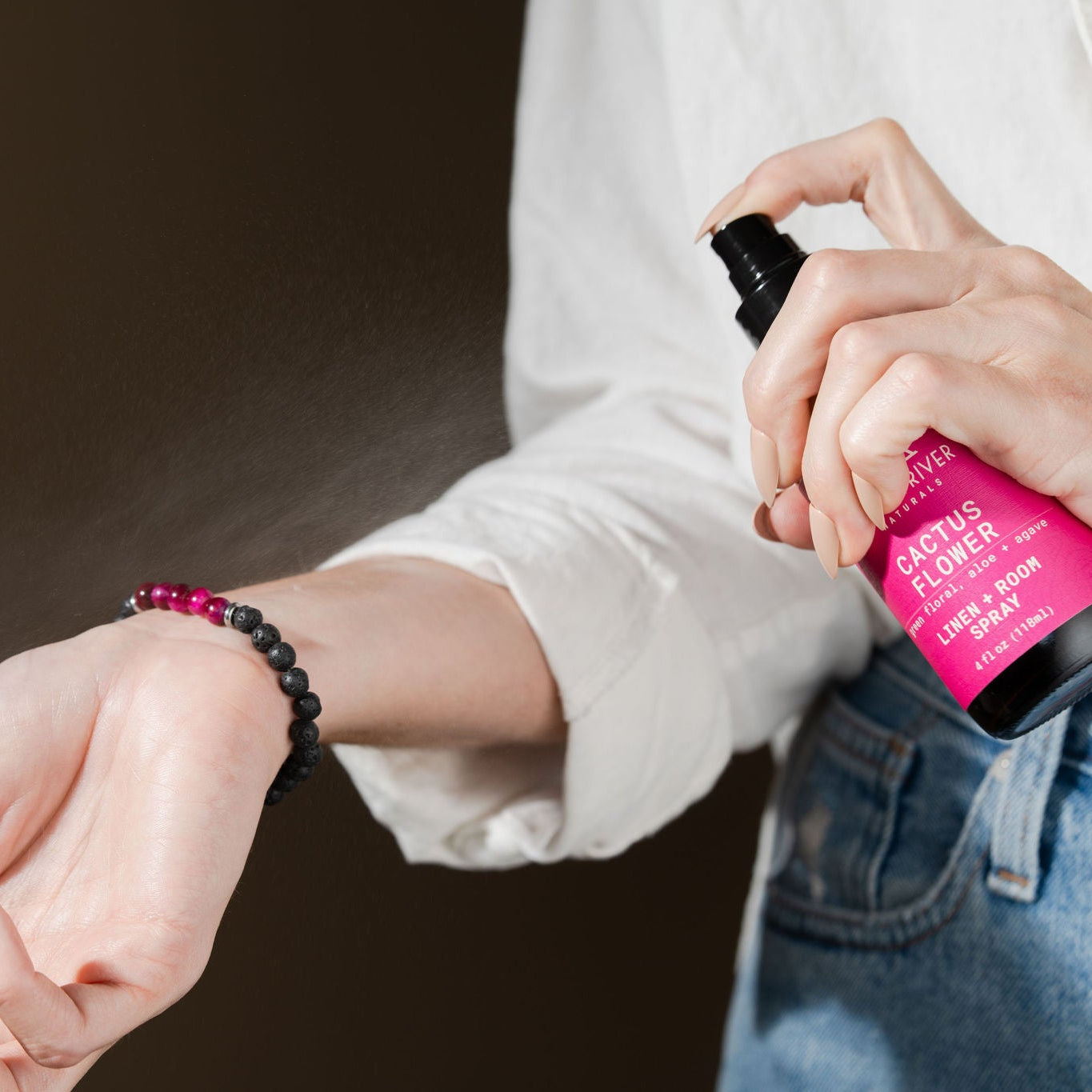 A person in a white shirt and jeans sprays cactus flower linen spray on their pink beaded bracelet
